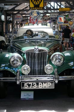 menu-03 Front view of a classic green MG TF 1953 on display in a vintage garage with retro signs.